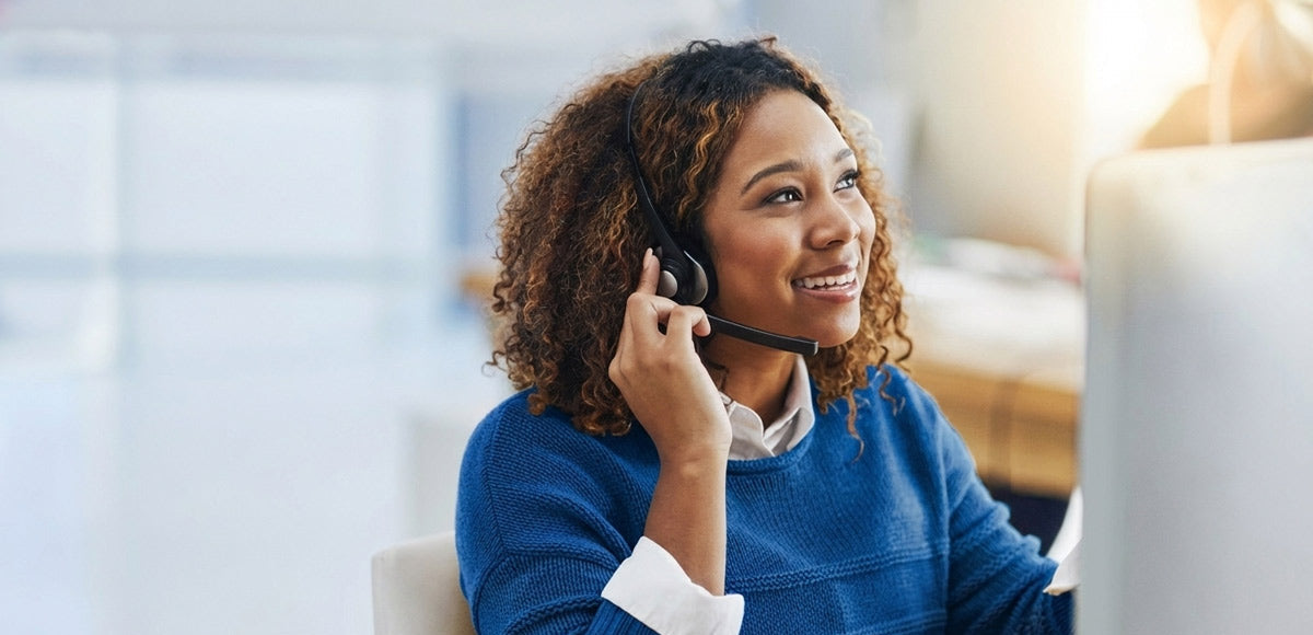 Woman wearing a headset and smiling, sitting at a desk with a computer.