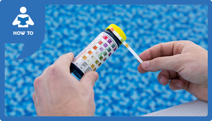 A hand holds a pool test strip next to a color chart over the pool water.
